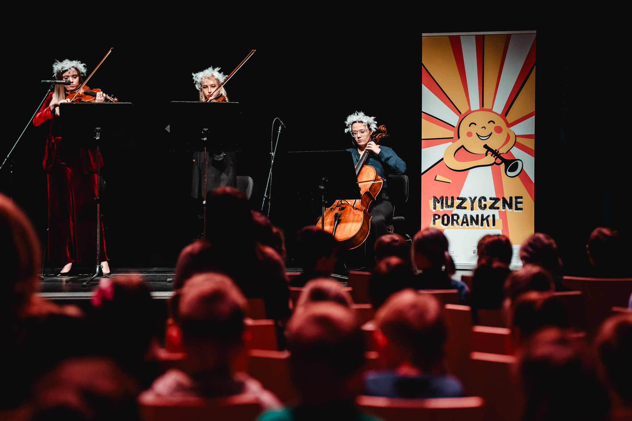 trzy kobiety muzyczki grające na instrumentach muzycznych na scenie. Na widowni siedzą dzieci.
