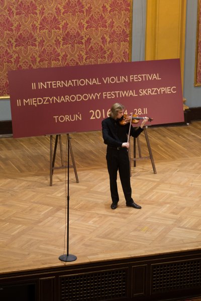 mężczyzna w czarnym stroju gra na skrzypcach na scenie, za nim baner z nazwą festiwalu