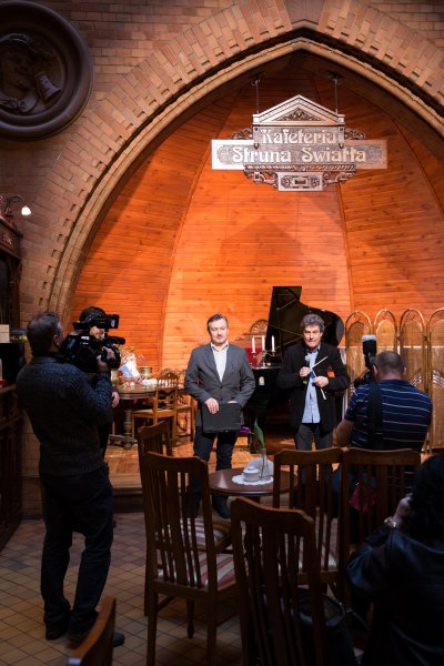 dwaj mężczyźni stoją na scenie w kafeterii, dziennikarze fotografują ich