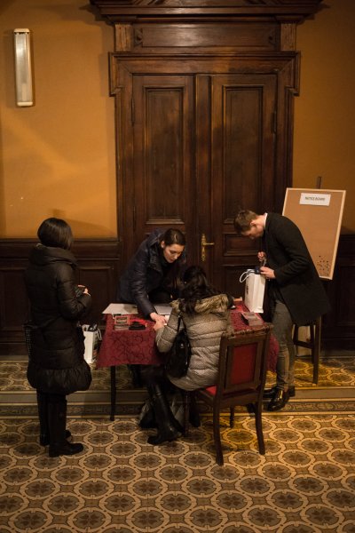 przy stole tyłem do obiektywu siedzi kobieta obok niej stoi kobieta w ciemnym płaszczu z drugiej strony pochylona kobieta nad stołem pokazuje coś dłonią obok kobiety pochylonej stoi mężczyzna z torbą papierową w dłoniach  