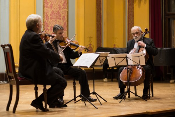na scenie trzech starszych mężczyzn siedzi grając na instrumentach smyczkowych