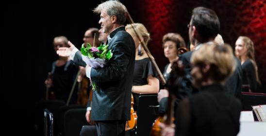 Solista koncertu stoi z kwiatami, za nim stoi orkiestra