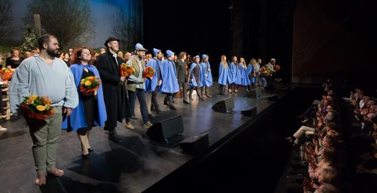 Chór flisaczy w niebieskich pelerynach i kapelusikach stoi z przodu sceny, soliści trzymają kwiaty, publiczność bije brawo.