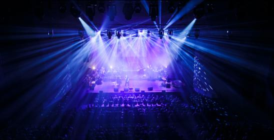 Widok sceny podczas koncertu z perspektywy, z miejsc balkonowych, wyciemniona widownia, mocne reflektorowe światło w oknie sceny