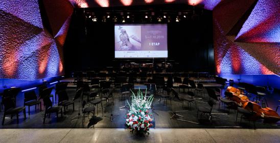 Sala koncertowa z ustawionymi krzesłami i pulpitami dla orkiestry, z przodu stoi bukiet kwiatów w koszyku a z tyłu na ekranie wyświetla się nazwa pierwszego etapu konkursu
