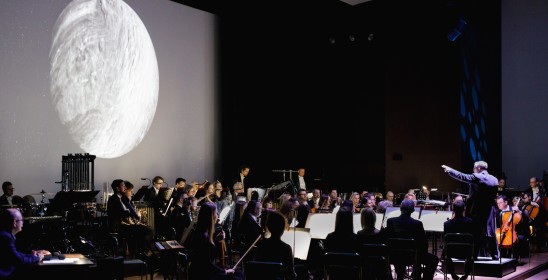 orkiestra na scenie na tle wyświetlanej planety