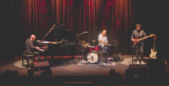 trzech mężczyzn grających na fortepianie, zestawie perkusyjnym i gitarze basowej