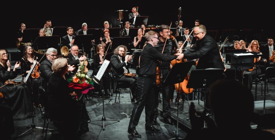 część orkiestry na scenie oraz trzech gratulujących sobie mężczyzn