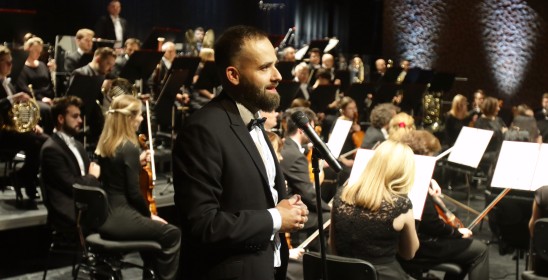 meżczyzna mówiący do mikrofonu, w tle siedzi orkiestra na scenie