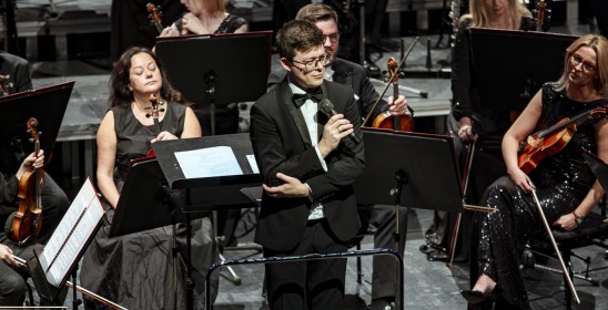 mężczyzna mówiący do mikrofonu, za nim siedzą kobiety