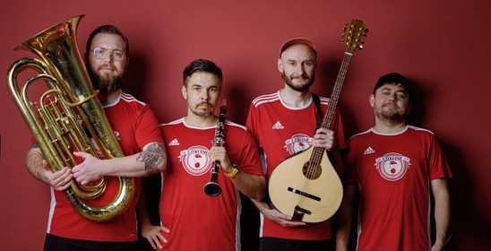 czterech mężczyzn w czerwonych koszulkach trzymajacych różne instrumenty muzyczne