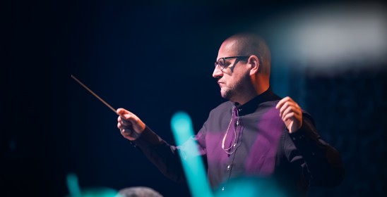 mężczyzna w czarnej koszuli trzyma ręce w górze, w prawej dłoni trzyma batutę