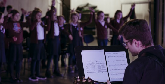 mężczyzna siedzący tyłem do fotografa grający na gitarze, przed nim pulpit z nutami a w tle skaczące dzieci