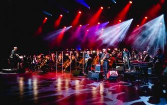 scena sali koncertowej w czerwonym oświetleniu, na scenie orkiestra symfoniczna z dyrygentem i zespół muzyczny Raz Dwa Trzy