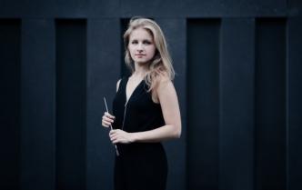 młda kobieta (Agata Zając) z blond włosami do ramion w czarnym stroju w dłoniach trzyma batutę drygencką