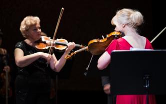 dwie kobiety stojąc na przeciw siebie grają na skrzypcach