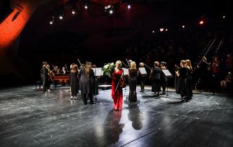 kobieta w czerwonej sukni z bukietem kwiatami schodzi ze sceny za nią stoją muzycy z instrumentami smyczkowymi, w tle widownia 
