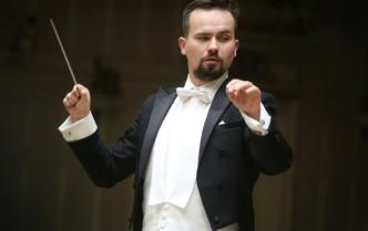 męzczyzna (Adam Banaszak) z brudką we fraku z uniesionymi rękami w prawej dłoni trzyma batutę drygencką