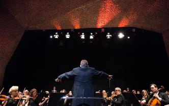 przed orkiestrą symfoniczą stoi mężczyzna we fraku z rozłożonymi rękoma - dyrygent