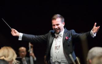 uśmiechnięty mężczyzna (dyrygent Adam Banaszak) z brudką w białej koszuli i ciemnym fraku stoi z rozłożonymi rękami w prawej dłoni trzyma batutę drygencką