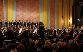 orkiestra siedzi na scenie, za nimi chóry, widok na głowy publiczności z tyłu