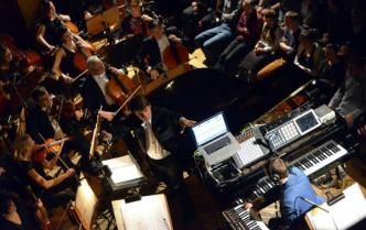 mężczyzna z dużą ilością sprzętu elektronicznego siedzi przy fortepianie, obok niego dyrygent we fraku, dalej grająca orkiestra