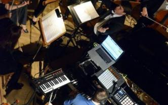 mężczyzna z dużą ilością sprzętu elektronicznego siedzi przy fortepianie, obok niego dyrygent we fraku, dalej grająca orkiestra