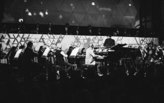 orkiestra siedzi na scenie, dalej DJe, za nimi projekcje wyświetlane na ścianie