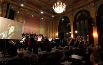 orkiestra siedzi na scenie, za nimi chóry, widok na głowy publiczności z tyłu, na ekranie z boku  wyświetlona twarz kobiety