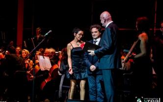 dwaj mężczyźni i kobieta w strojach wieczorowych stoją na scenie, za nimi orkiestra
