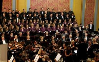 od góry chór mężczyźni w czarnych garniturach niżej kobiety w fioletowych sukienkach, niżej orkiestra siedzi na scenie