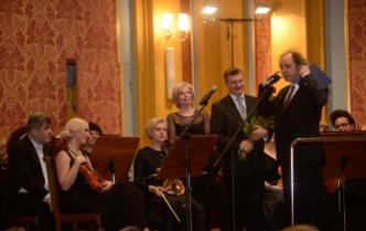 mężczyzna z kwiatami przemawia do mikrofonu, inne osoby przyglądają się