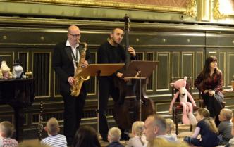mężczyźni grają na saksofonie oraz kontrabasie, przed nimi siedzą dzieci