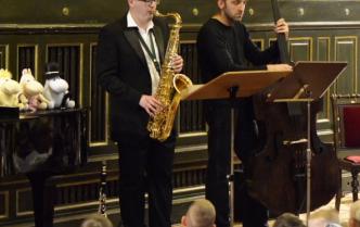 mężczyźni grają na saksofonie oraz kontrabasie, przed nimi siedzą dzieci, na fortepianie obok leżą maskotki muminków