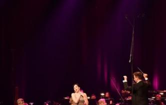 śpiewająca sopranistka ze złożonymi rękoma na lewym ramieniu  z prawej strony stojący bokiem z rękoma w górze stoi dyrygent  przed nim część orkiestry kobiety i mężczyźni grający na instrumentach smyczkowych