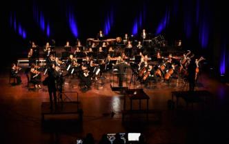 Na scenie orkiestra kobiety i mężczyźni grający na instrumentach oraz przed orkiestrą na podeście dyrygent - Dainius Pavilionis
