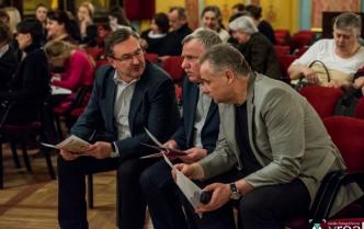 trzej mężczyzni rozmawiają siedząc w fotelach w sali wielkiej Dworu Artusa