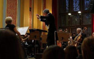 dyrygent z wyciągniętymi dłońmi w tle orkiestra