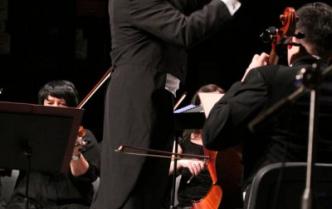 dyrygent na podeście wykonujący gest ręką, obok mężczyzna z wiolonczelą siedzący tyłem