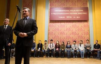 przy mikrofonie stoi dyrektor Toruńskiej Orkiestry Symfonicznej , z lewej strony stoi mężczyzna prowadzący konkurs skrzypcowy w tle uczestnicy konkursu skrzypcowego