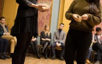dwie kobiety stoja jedna trzyma misę do losowania druga z długimi ciemnymi włosami w dłoni trzyma wylosowaną kartkę