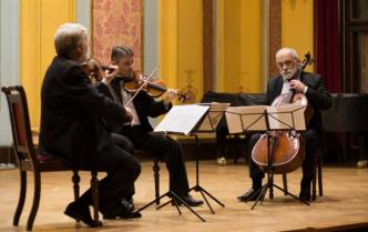 na scenie trzech starszych mężczyzn siedzi grając na instrumentach smyczkowych