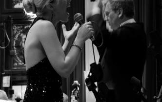 czarno-białe zdjęcie śpiewającej blond kobiety, w tle dyrygent