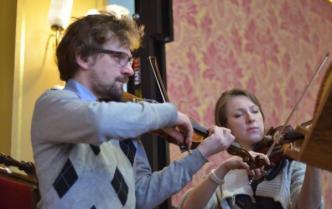 mężczyzna w swetrze grający na skrzypcach, w tle grająca kobieta