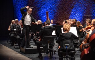 na scenie stoi mężczyzna w okularach w czarnym fraku-dyrygent w uniesionej prawej ręce trzyma batutę dyrygencką przed nim w półkolu siedzą muzycy orkiestry symfonicznej 