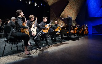na scenie kobieta i trzech mężczyzn siedzą z gitarami, w dali po prawej stroni męzczyzna-dyrygent z uniesionymi rękami za nim muzycy graja na wiolonczelach i kontrabasie 