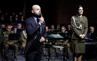 mężczyzna w garniturze mówiący do mikrofony i stojąca kobieta w mundurze