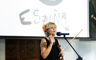 kobieta w czarnym stroju z kwiatem we włosach, trzymając skrzypce przemawia do mikrofonu na tle prezentacji z nazwą kraju - Espana