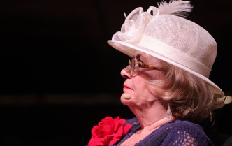 zbliżenie na twarz starszej kobiety w granatowej sukni z czerwonym kwiatem i w białym kapeluszu