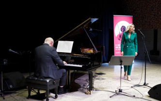mężczyzna grający przy fortepianie a obok niego stojąca i śpiewająca kobieta w zielonym garniturze